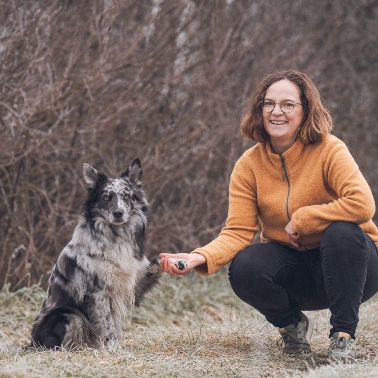 Eine lächelnde Person in orangefarbener Jacke sitzt neben einem Hund im Freien.