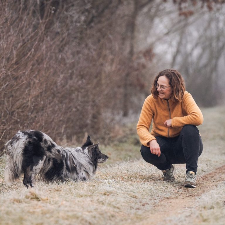 Ein Mensch in orangefarbener Jacke kniet neben einem Hund auf einem verschneiten Weg.